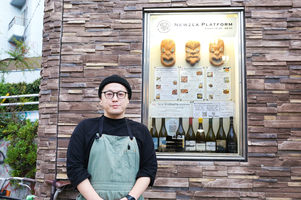 お店の看板の前に経つ箸本オーナーシェフ